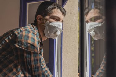 Coronavirus. Evde karantina var. Corona virüsünün hasta adamı pencereden dışarı bakıyor ve maske takıyor ve evdeki hastalıktan korunuyor.. 