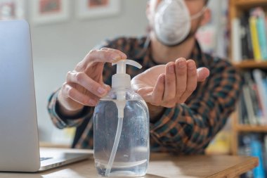 Coronavirus. Koruyucu maske takan bir adam evde çalışıyor. Koronavirüs için koruyucu maske takarak karantina. Evden çalışıyorum. Ellerini dezenfektan jeliyle temizliyor. Termometre ateşi denetimi.