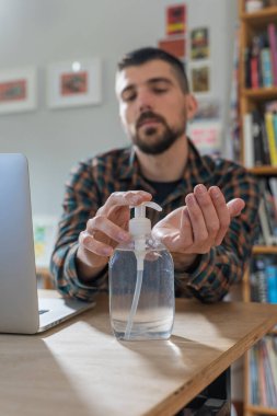 Coronavirus. Koruyucu maske takan bir adam evde çalışıyor. Koronavirüs için koruyucu maske takarak karantina. Evden çalışıyorum. Ellerini dezenfektan jeliyle temizliyor. Termometre ateşi denetimi.
