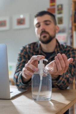 Coronavirus. Koruyucu maske takan bir adam evde çalışıyor. Koronavirüs için koruyucu maske takarak karantina. Evden çalışıyorum. Ellerini dezenfektan jeliyle temizliyor. Termometre ateşi denetimi.