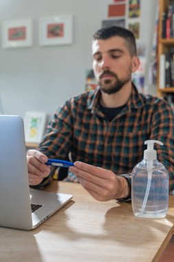 Coronavirus. Koruyucu maske takan bir adam evde çalışıyor. Koronavirüs için koruyucu maske takarak karantina. Evden çalışıyorum. Ellerini dezenfektan jeliyle temizliyor. Termometre ateşi denetimi.
