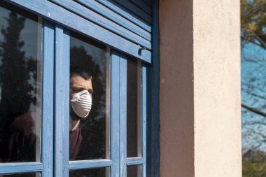 Coronavirus. Corona virüsünün hasta adamı pencereden dışarı bakıyor ve maske takıyor ve evdeki hastalıktan korunuyor. Karantinaya alın. Hasta enfeksiyonu önlemek için izole edilmiş. Pandemia. Ev.