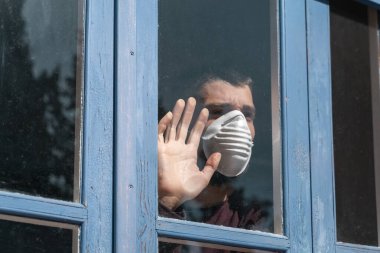 Coronavirus. Corona virüsünün hasta adamı pencereden dışarı bakıyor ve maske takıyor ve evdeki hastalıktan korunuyor. Karantinaya alın. Hasta enfeksiyonu önlemek için izole edilmiş. Pandemia. Ev.