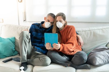 Coronavirus. Evde karantinada kanepede oturup televizyon izleyen bir çift. Kanepede oturan çift bulaşıcı hastalıktan korunmak için maske takıyor. Evde kal.. 