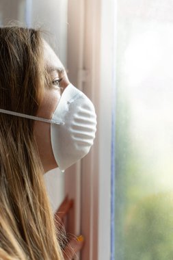 Coronavirus. Corona virüsünün hasta kadını pencereden dışarı bakıyor ve maske takıyor ve evdeki hastalıktan korunuyor. Karantinaya alın. Hasta enfeksiyonu önlemek için izole edilmiş. Ev.