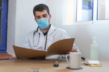 Coronavirus. Doktor teşhis üzerinde çalışıyor. Coronavirüs salgını sırasında reçeteyi hastanenin masasına yazıyor. Önlem için yüz maskesi ve dezenfektan jeli. Sağlık çalışanları. Coronavirus testi. 