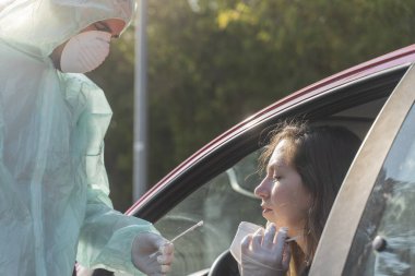 Coronavirus. Tam korumalı ekipmanlı bir polis teknisyeni arabasında oturan genç bir kadından örnek alıyor. Coronavirus mobil test ünitesi. İzole edilmiş. Karantina. 