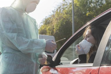 Coronavirus. Tam korumalı ekipmanlı bir polis teknisyeni arabasında oturan genç bir kadından örnek alıyor. Coronavirus mobil test ünitesi. İzole edilmiş. Karantina. 