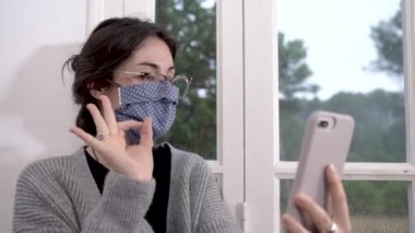 Coronavirus. Koronavirüs yüzünden karantinada olan bir kadın koruyucu maske takıyor, evden çalışıyor ve koltukta cep telefonu kullanıyor. Görüntülü arama. Cep telefonuyla video konferansı.