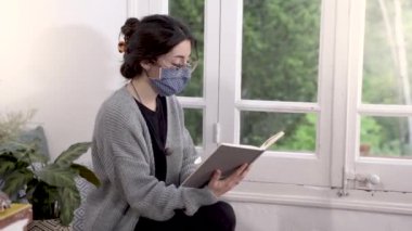 Coronavirus. Corona virüsü yüzünden karantinada olan koruyucu maske takan bir kadın kitap okuyor. Evde sakin ol. Ev kilitlenmesi