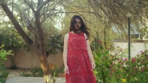 Schöne Mädchen im roten Kleid spaziert allein durch den grünen Garten — Stockvideo