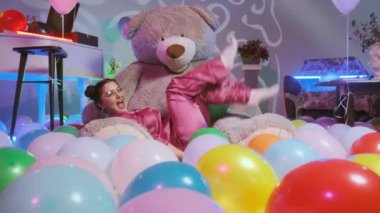 O da pembe pijama giyip oyuncak ayının üzerine oturman ve tatlı gibi davranman. Kameraya bak ve gülümse. Yerde çok renkli hava balonları, renkli neon ışıklarla süslenmiş bir oda.