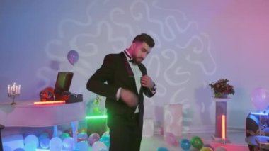 Siyah smokin giymiş yakışıklı bir adam kameraya bakıyor. Yerde çok renkli hava balonları, renkli neon ışıklarla süslenmiş bir oda.