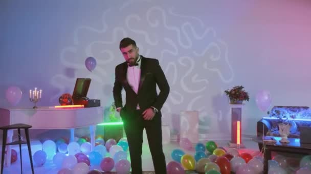 Hombre guapo que está en esmoquin negro mirando a la cámara posando, globos de aire multicolor en el suelo, habitación con decorado con luces de neón de colores — Vídeo de stock