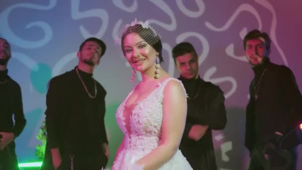 Braut blickt in die Kamera und spielt mit ihrem Schleier, Hintergrund mit Männern in Schwarz, Schönes Model-Mädchen im weißen Hochzeitskleid, Frau mit Frisur — Stockvideo