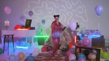 Pembe pijamalı bir kız elinde mavi bir balon, yerde çok renkli hava balonları, renkli neon ışıklarla süslenmiş bir oda.