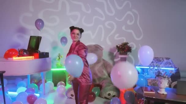 Девушка танцует в розовой пижаме есть синий шар у ее руки, разноцветные воздушные шары на полу, комната с украшенной красочными неоновыми огнями — стоковое видео