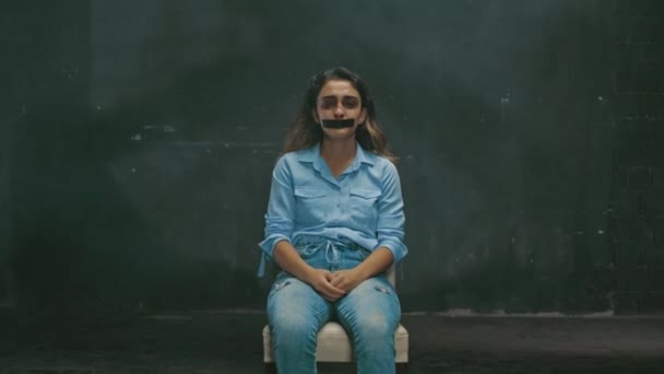 Μια νεαρή γυναίκα με μελανιές και αίμα στο πρόσωπό της. Ενδοοικογενειακή βία, κοιτάζοντας την κάμερα και κλαίγοντας, το στόμα είναι καλυμμένο με μαύρη ταινία — Αρχείο Βίντεο
