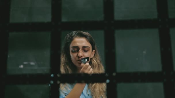 一个脸上有伤痕和血迹的年轻女人家庭暴力，看着镜头和哭泣，从嘴里掏出黑色带子，关在牢里的铁窗后面 — 图库视频影像