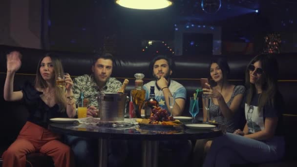 Eine Gruppe verschiedener Freunde, die miteinander kommunizieren, sitzt am Tisch der Disco-Bar mit bunten Getränken. Junge Leute genießen Entspannung, aromatischen Rauch, leckere Cocktails im Nachtclub — Stockvideo