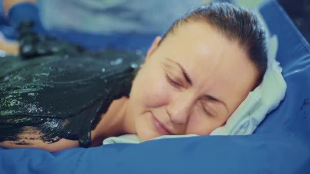 Mujer joven y relajada tumbada en la cama de spa para masajes corporales usando barro y especias por un terapeuta de masaje en un resort de lujo . — Vídeo de stock