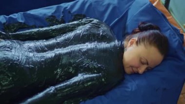 Rahatlamış genç bir kadın çamur kullanarak vücut masajı için spa yatağında yatıyor.