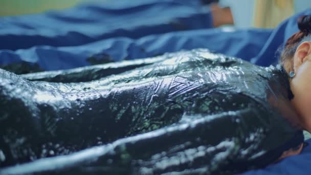 Uvolněná mladá žena ležící na lázeňské posteli pro masáž těla bahnem — Stock video