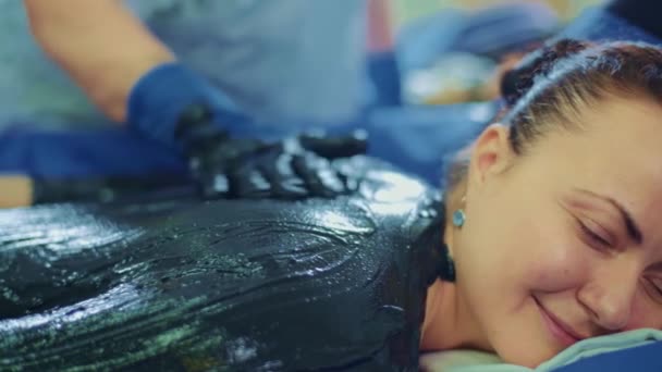 Mujer joven y relajada tumbada en la cama de spa para masajes corporales con barro y especias por un terapeuta de masaje en un spa de lujo. — Vídeos de Stock