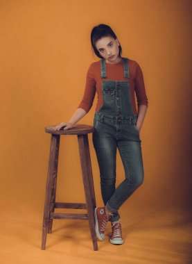 Kırmızı kazaklı ve mavi tulumlu güzel bir kız model. Sandalyenin yanında duruyor.,