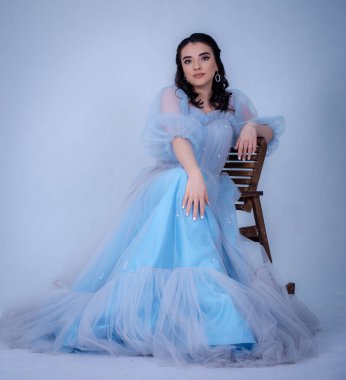 Prenses suretinde genç ve güzel bir manken. Mavi bir elbise giymiş, sandalyede oturuyor.,