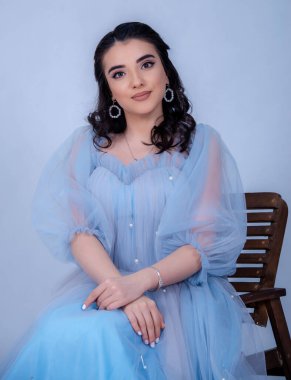 Prenses suretinde genç ve güzel bir manken. Mavi bir elbise giymiş, sandalyede oturuyor.,