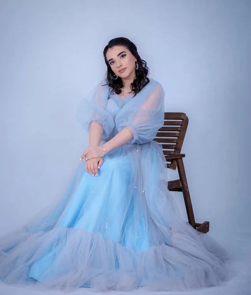 Prenses suretinde genç ve güzel bir manken. Mavi bir elbise giymiş, sandalyede oturuyor.,