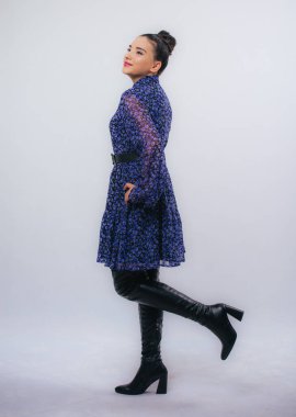 Stüdyoda poz veren mutlu mavi elbiseli güzel kadın beyaz arka planda izole edilmiş, ayaklarında siyah uzun çizmeler, tam boy portre.,