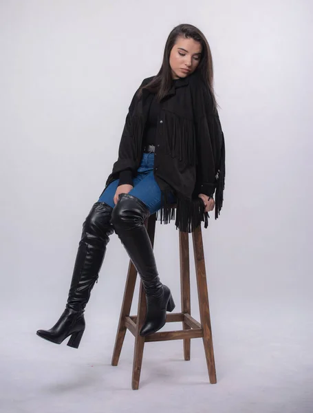 穿着黑色衣服的漂亮女模 穿着蓝色牛仔裤和黑色靴子的少女 坐在椅子上 站在工作室里 与白色背景隔离 — 图库照片