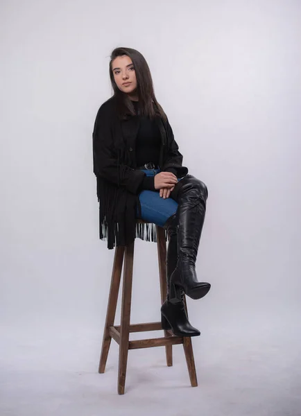 Beyaz arka planda izole edilmiş siyah elbiseli mutlu güzel kız model, mavi kot ve siyah çizmeli genç kız tam boy portre, bir sandalyede oturuyor.,