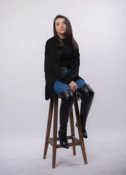 Beyaz arka planda izole edilmiş siyah elbiseli mutlu güzel kız model, mavi kot ve siyah çizmeli genç kız tam boy portre, bir sandalyede oturuyor.,