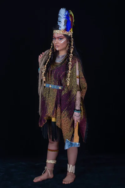 アメリカ先住民 スタジオでポーズをとる伝統的なドレスのインド人女性 インドの羽の帽子とカラフルなメイクを持つシャーマン女性の肖像画 彼の手に斧を保持 — ストック写真