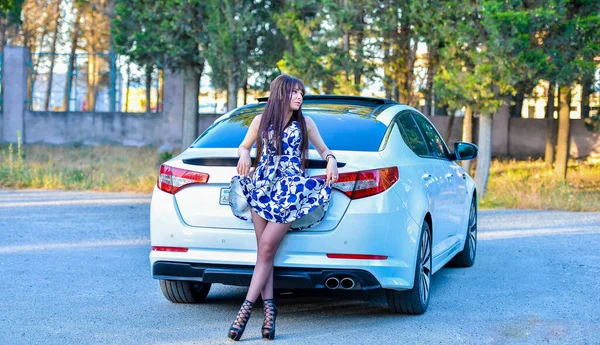 Genç, beyaz bir kadın, uzun düz saçlı, orman manzarasında beyaz spor arabanın yanında duruyor ve poz veriyor. Açık hava moda portresi,
