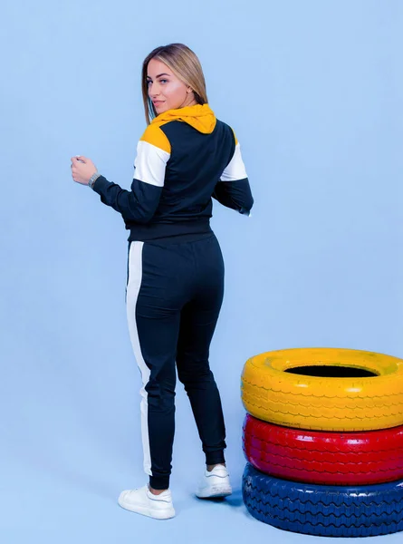 Spor giyimli güzel sarışın manken kız mavi arka planda çok renkli lastiklerin yanında poz veriyor.,