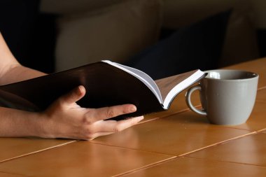 Evde bir fincan kahveyle kitap okuyan genç bir kadın.