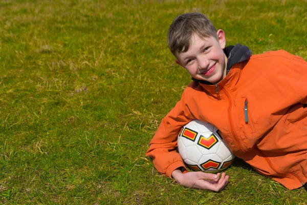 Chłopiec Siwymi Włosami Pomarańczowej Kurtce Leży Trawniku Piłką Rękach — Zdjęcie stockowe