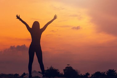 Kadın silueti gün batımında arka planda, mutlu kız ellerini kaldırıp gökyüzüne bakıyor..