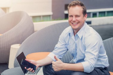 Gündelik tarzı olan genç, mutlu, beyaz bir adam bilgisayarda çalışıyor..