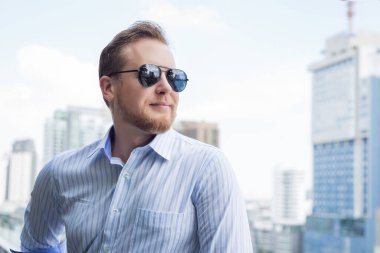 Beyaz güneş gözlüğü takan genç bir adam yüksek binanın çatısında şehir manzarasına bakıyor..