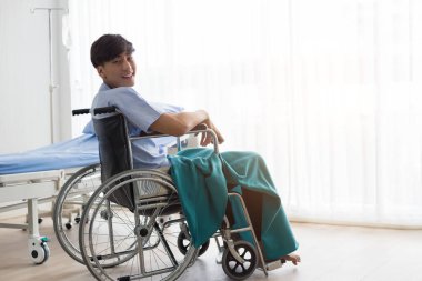 Hastanede tekerlekli sandalyede oturan gülümseyen yüzlü genç Asyalı adam..