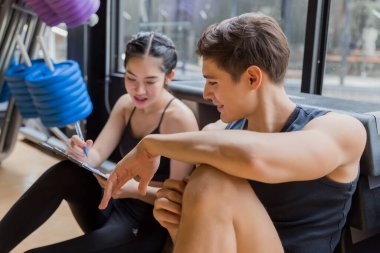 Genç ırklar arası bir çift spor salonunda etkinlik planına bakıyor ve sohbet ediyorlar..