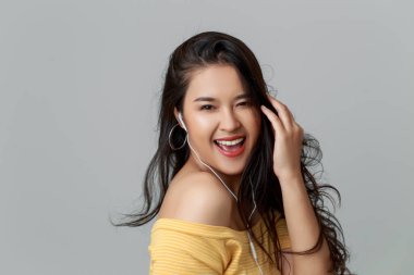 Close up young attractive Asian woman wearing yellow t shirt, earphones listening music with smiley face, studio head shot isolated on grey background. clipart