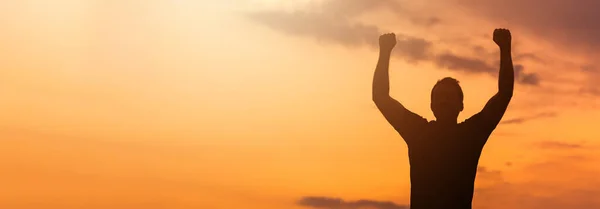 Silhouette of a man with hands raised in the sunset, empowered, victory concept. Banner ratio.