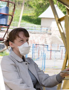 Güneşli bir bahar günü, şehir parkında salıncakta salıncakta oturan tıbbi solunum cihazlı genç bir kadın. Coronavirus koruması. Açık havada..