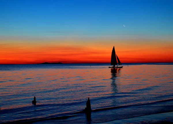 Backlit sailboat sailing on a sunset. The photo was taken on a beach in Uruguay. There are two remains of pilings that form an imaginary triangle with the sailboat. Its shadow is reflected in the waters. There are two waves and an island.